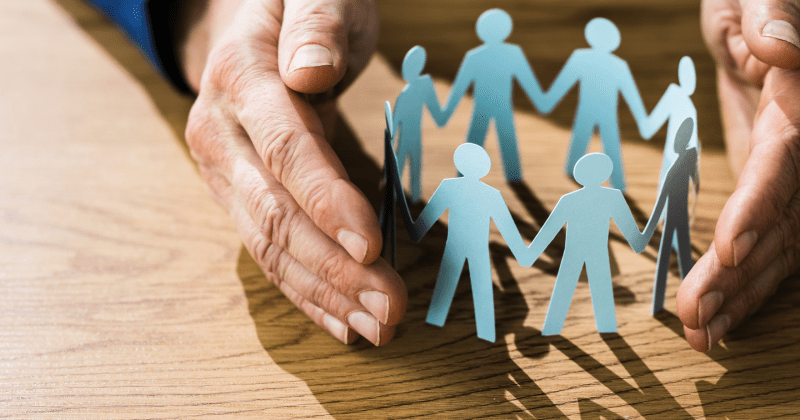 An elderly person's hands lightly wrap around a chain of paper figures in the shape of people. It represents adult safeguarding.